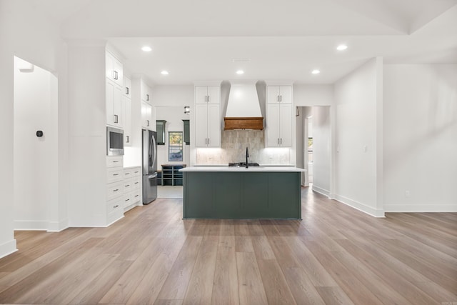 kitchen with light wood-type flooring, premium range hood, stainless steel appliances, a center island with sink, and white cabinetry