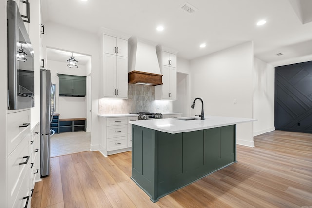 kitchen with sink, premium range hood, light hardwood / wood-style floors, a center island with sink, and white cabinets