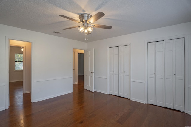 unfurnished bedroom with a textured ceiling, two closets, ceiling fan, and dark hardwood / wood-style floors