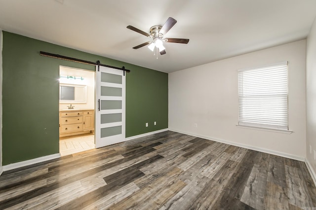 unfurnished bedroom featuring ceiling fan, a barn door, dark hardwood / wood-style flooring, and connected bathroom