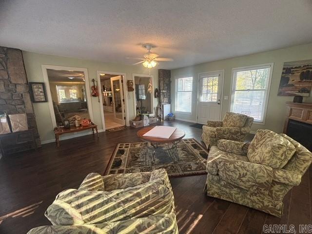 living room with a textured ceiling, a stone fireplace, ceiling fan, and dark wood-type flooring