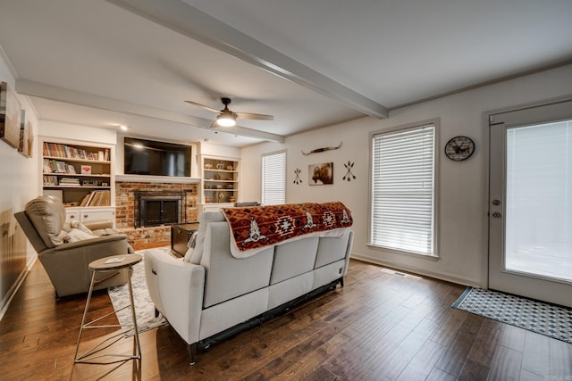 living room with built in shelves, ceiling fan, dark wood-type flooring, a brick fireplace, and beamed ceiling