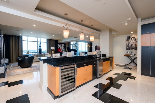 kitchen with light brown cabinetry, sink, wine cooler, hanging light fixtures, and an island with sink