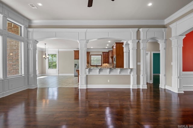 unfurnished living room featuring ceiling fan with notable chandelier, decorative columns, crown molding, and dark wood-type flooring