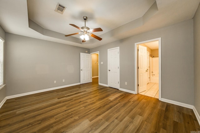 unfurnished bedroom with a tray ceiling, ensuite bath, ceiling fan, and dark wood-type flooring