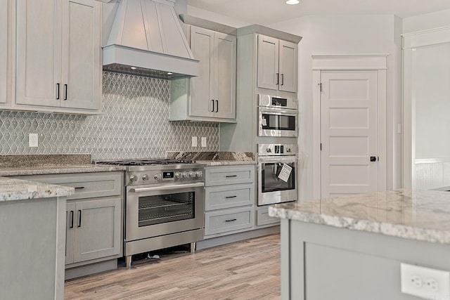 kitchen featuring gray cabinetry, stainless steel appliances, light stone counters, light hardwood / wood-style flooring, and custom range hood