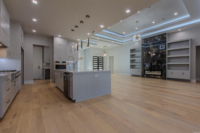 kitchen with a raised ceiling, a spacious island, hanging light fixtures, light wood-type flooring, and stainless steel appliances