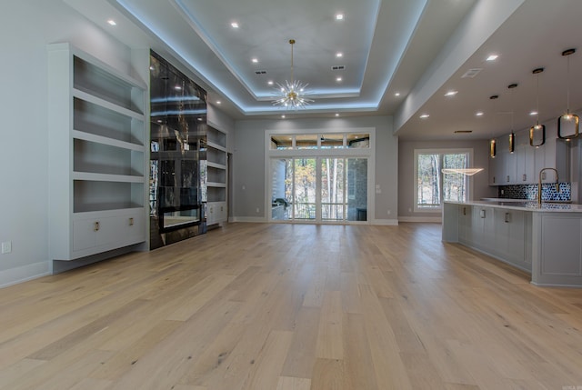 unfurnished living room featuring a raised ceiling, sink, built in features, light hardwood / wood-style floors, and a chandelier