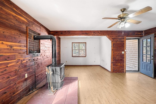 unfurnished living room with ceiling fan, wooden walls, wood finished floors, baseboards, and a wood stove