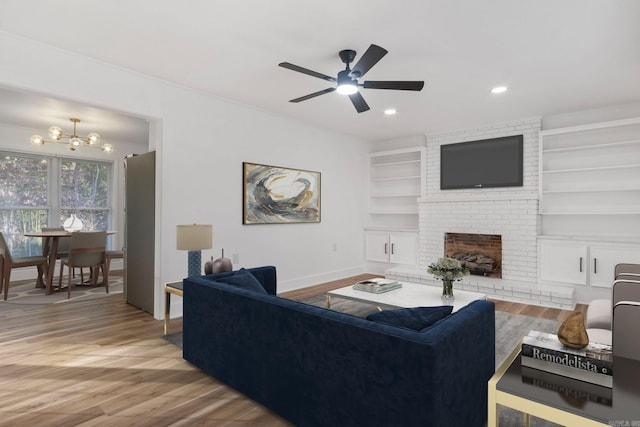 living room featuring built in shelves, light hardwood / wood-style floors, ceiling fan with notable chandelier, and a brick fireplace