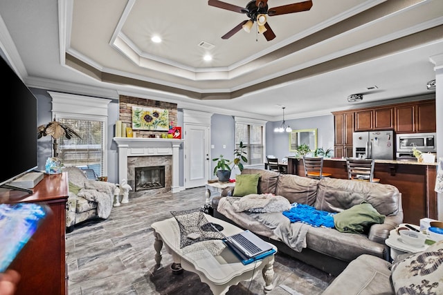 living room featuring a tray ceiling, crown molding, a fireplace, and ceiling fan with notable chandelier