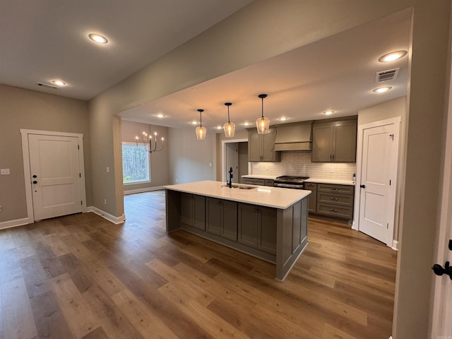 kitchen with pendant lighting, gray cabinets, premium range hood, a kitchen island with sink, and stainless steel range oven