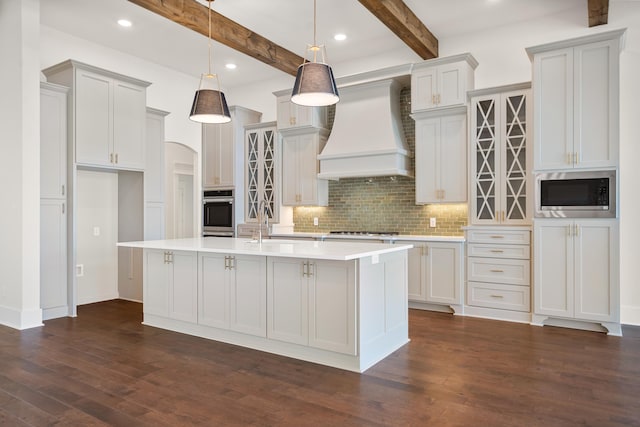 kitchen featuring appliances with stainless steel finishes, custom range hood, a kitchen island with sink, and decorative light fixtures