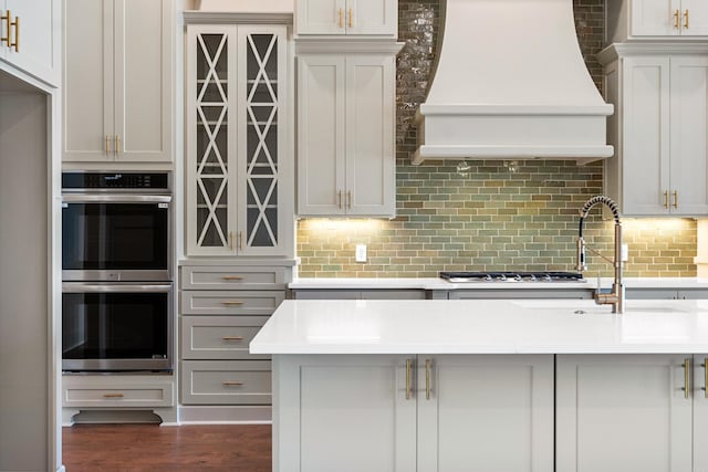 kitchen with dark wood-type flooring, sink, custom range hood, stainless steel double oven, and backsplash