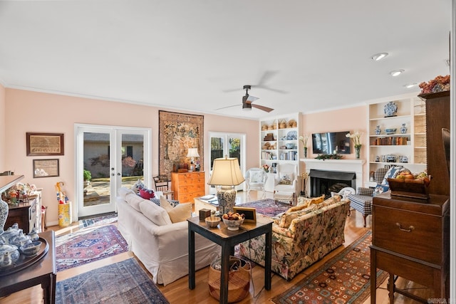 living room featuring built in shelves, ceiling fan, french doors, light hardwood / wood-style floors, and ornamental molding