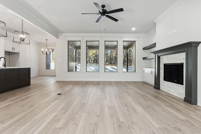 unfurnished living room with ceiling fan with notable chandelier, a stone fireplace, sink, light wood-type flooring, and ornamental molding