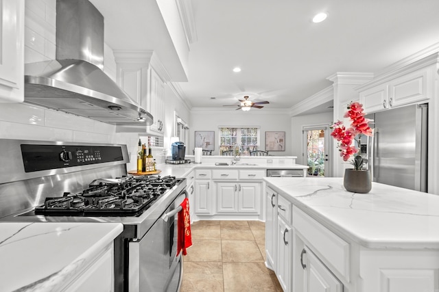 kitchen with light stone countertops, wall chimney range hood, crown molding, white cabinets, and appliances with stainless steel finishes