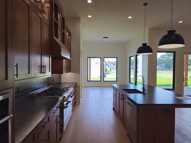 kitchen featuring sink, hanging light fixtures, stainless steel appliances, an island with sink, and light wood-type flooring