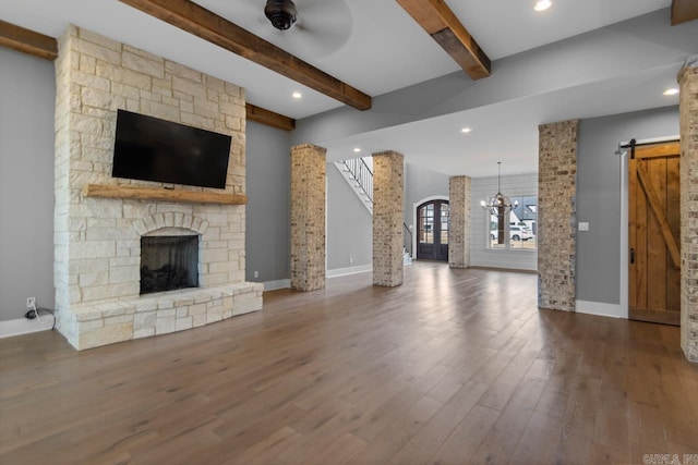 unfurnished living room with ceiling fan with notable chandelier, a barn door, a fireplace, beamed ceiling, and wood-type flooring