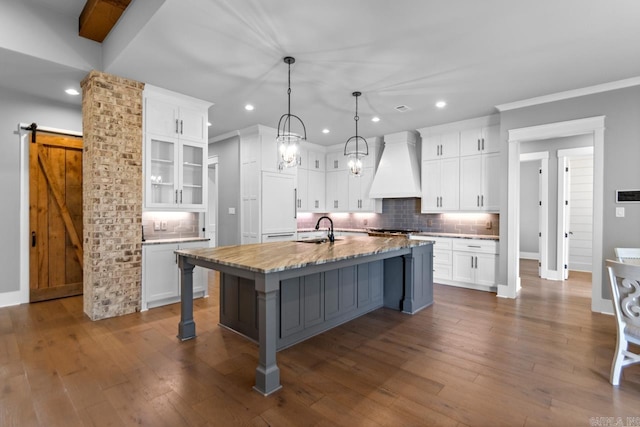 kitchen with custom exhaust hood, backsplash, white cabinets, a barn door, and an island with sink