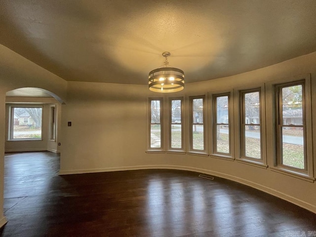 empty room with arched walkways, dark wood-style flooring, a notable chandelier, visible vents, and baseboards