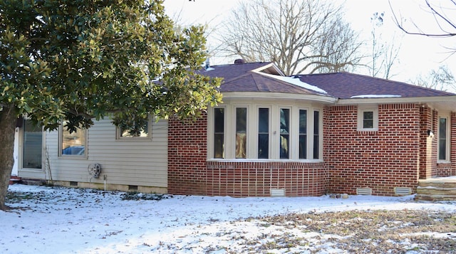 snow covered property with roof with shingles, brick siding, and crawl space