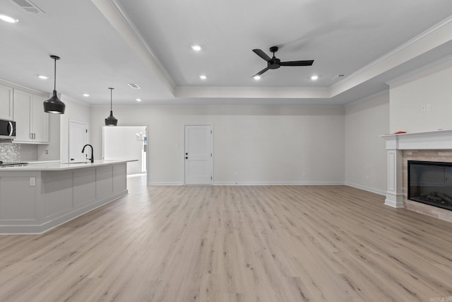 unfurnished living room with ceiling fan, sink, a raised ceiling, light hardwood / wood-style flooring, and a tiled fireplace