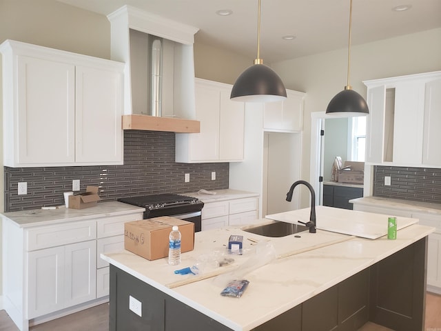 kitchen with light stone countertops, wall chimney exhaust hood, pendant lighting, a kitchen island with sink, and white cabinets