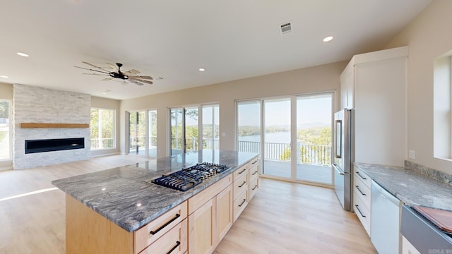 kitchen featuring light brown cabinetry, stainless steel appliances, a water view, dark stone countertops, and a fireplace