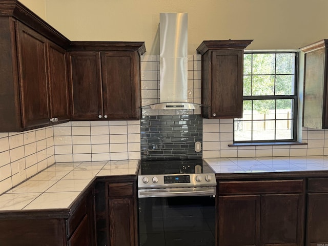 kitchen with tile countertops, stainless steel electric range, backsplash, wall chimney exhaust hood, and dark brown cabinets