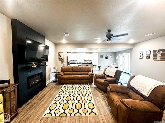 living room featuring ceiling fan, a large fireplace, a textured ceiling, and light hardwood / wood-style flooring