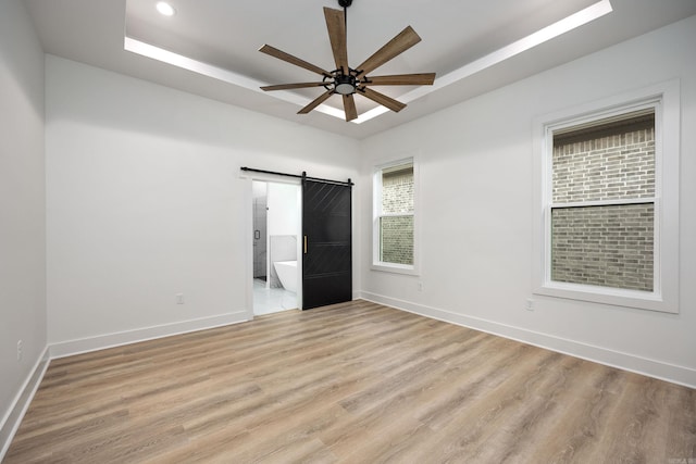 unfurnished bedroom featuring a barn door, a tray ceiling, ceiling fan, and light hardwood / wood-style floors