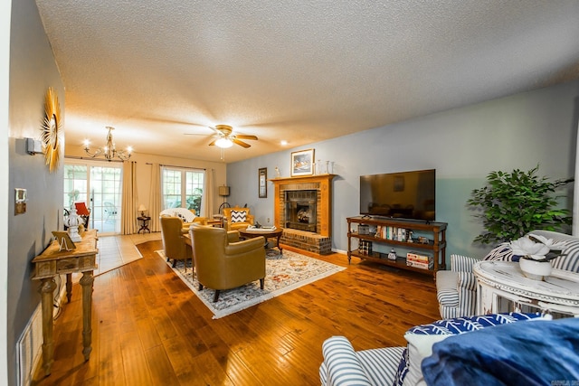 living room featuring hardwood / wood-style flooring, ceiling fan with notable chandelier, a textured ceiling, and a brick fireplace