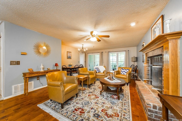 living room with ceiling fan with notable chandelier, hardwood / wood-style floors, a textured ceiling, and a brick fireplace