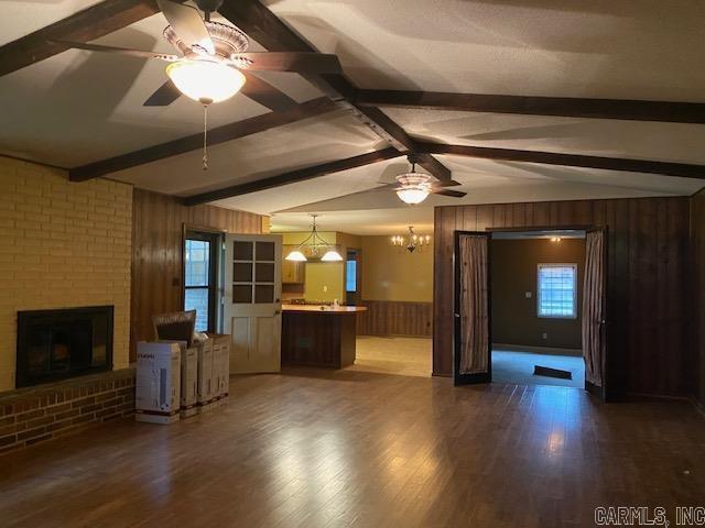 unfurnished living room with a brick fireplace, lofted ceiling with beams, dark hardwood / wood-style floors, wooden walls, and ceiling fan with notable chandelier