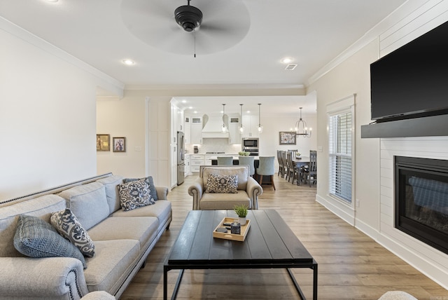 living area featuring crown molding, visible vents, light wood-style flooring, a glass covered fireplace, and ceiling fan with notable chandelier