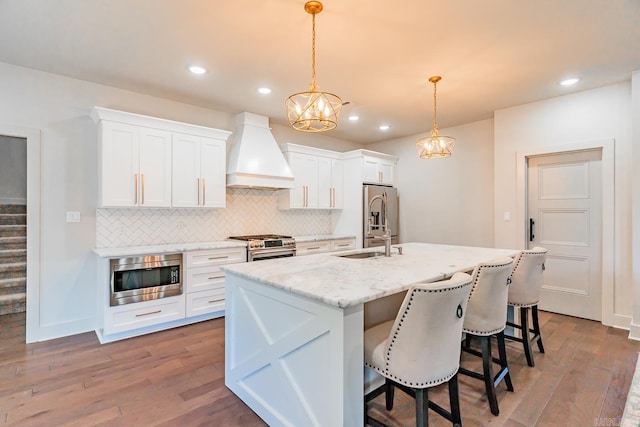 kitchen featuring pendant lighting, premium range hood, white cabinets, a center island with sink, and stainless steel appliances