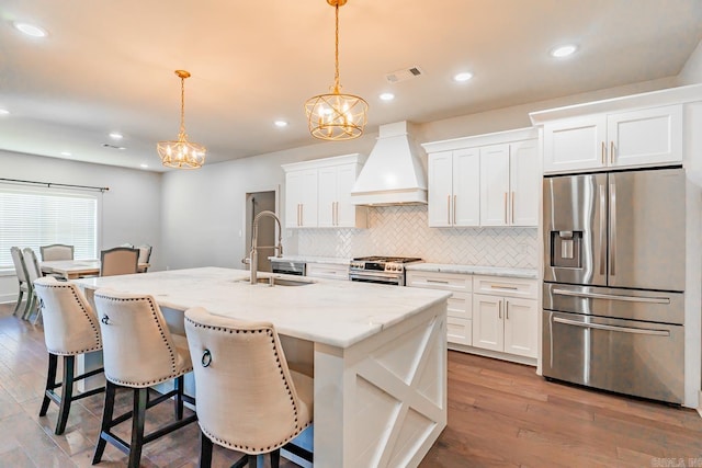 kitchen featuring appliances with stainless steel finishes, premium range hood, a kitchen island with sink, sink, and hanging light fixtures