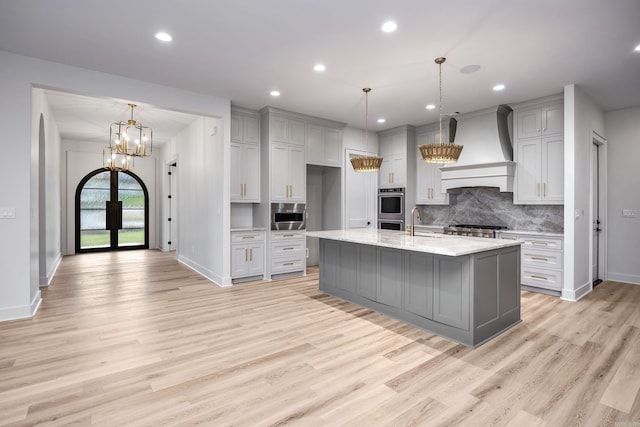 kitchen with appliances with stainless steel finishes, premium range hood, pendant lighting, gray cabinets, and an island with sink