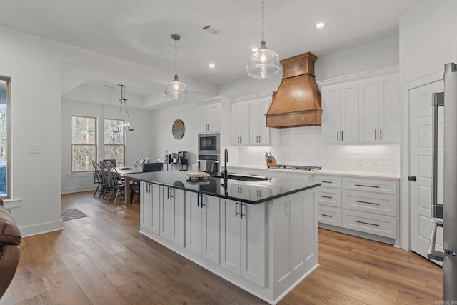 kitchen with white cabinetry, stainless steel appliances, an island with sink, decorative light fixtures, and custom exhaust hood