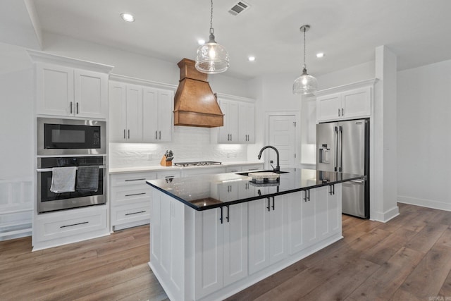 kitchen with a kitchen island with sink, premium range hood, sink, appliances with stainless steel finishes, and white cabinetry