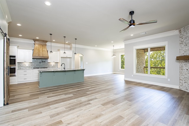 kitchen with white cabinets, premium range hood, hanging light fixtures, and an island with sink