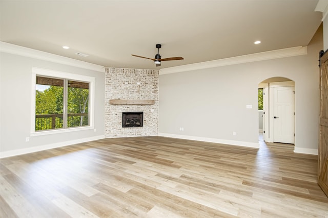 unfurnished living room with ornamental molding, ceiling fan, a barn door, light hardwood / wood-style floors, and a stone fireplace