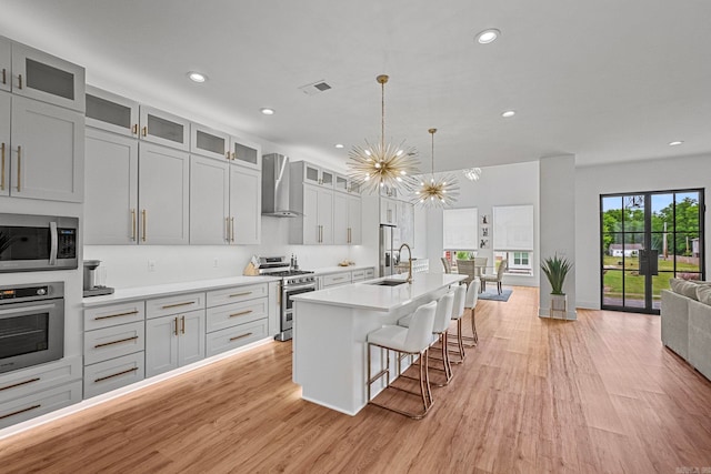 kitchen featuring an inviting chandelier, a center island with sink, hanging light fixtures, wall chimney exhaust hood, and appliances with stainless steel finishes