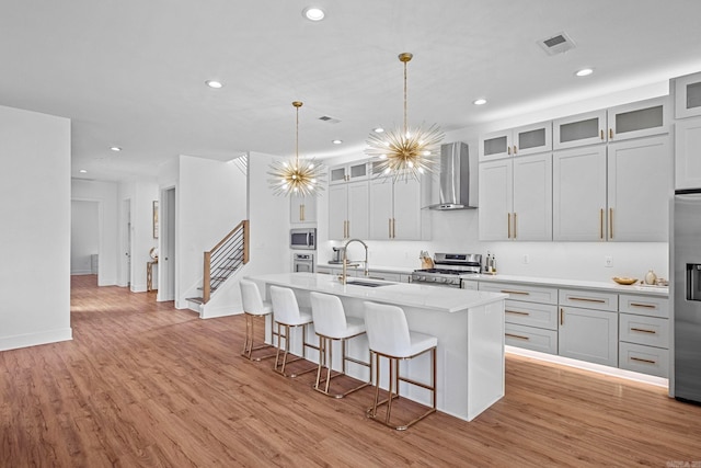 kitchen featuring appliances with stainless steel finishes, a kitchen island with sink, sink, wall chimney range hood, and hanging light fixtures