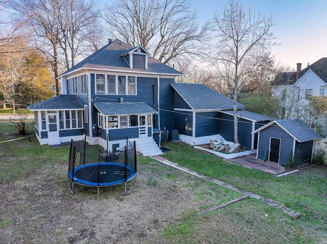 back house at dusk featuring a lawn, a sunroom, an outdoor structure, a trampoline, and central air condition unit