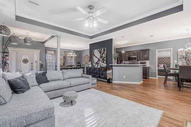 living room featuring ornate columns, ornamental molding, ceiling fan with notable chandelier, a tray ceiling, and light hardwood / wood-style flooring