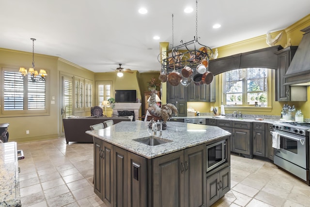 kitchen with light stone countertops, ceiling fan with notable chandelier, stainless steel appliances, dark brown cabinets, and a center island with sink