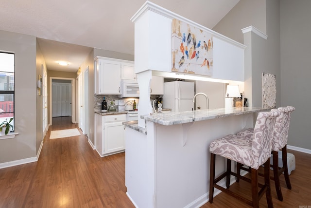 kitchen with white appliances, a kitchen breakfast bar, kitchen peninsula, tasteful backsplash, and white cabinetry