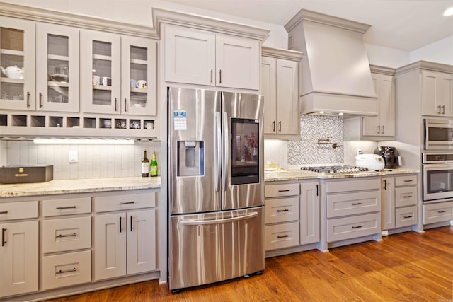 kitchen with light stone countertops, backsplash, custom range hood, stainless steel appliances, and wood-type flooring